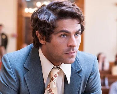 A man in a courtroom setting wearing a blue suit, white shirt, and patterned tie, looking intently forward.