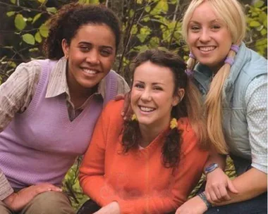 Three young women sit together smiling outdoors, surrounded by trees and greenery.