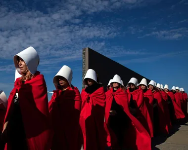 Protesters dressed as handmaids in red cloaks and white bonnets march under a cloudy blue sky.