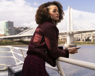 Woman in red jacket with curly hair stands on a boat, holding a phone, with a modern bridge in the background.