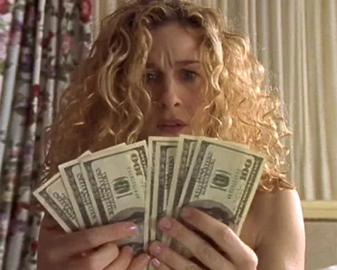 Woman with curly hair holds several $100 bills, looking surprised.