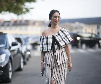 A woman wearing an off-shoulder, checkered top and striped skirt, walking on a street with parked cars in the background.