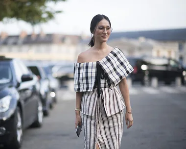 A woman wearing an off-shoulder, checkered top and striped skirt, walking on a street with parked cars in the background.