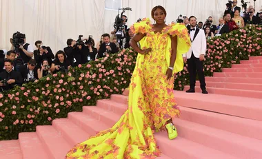 Individual in a yellow gown with pink flowers poses on the Met Gala 2019 red carpet, surrounded by photographers.