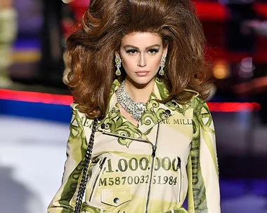 Model on runway wearing a dress with dollar bill print and large hairstyle, adorned with statement jewelry.