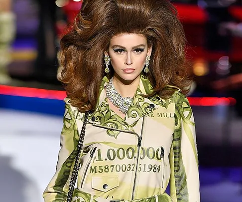 Model on runway wearing a dress with dollar bill print and large hairstyle, adorned with statement jewelry.