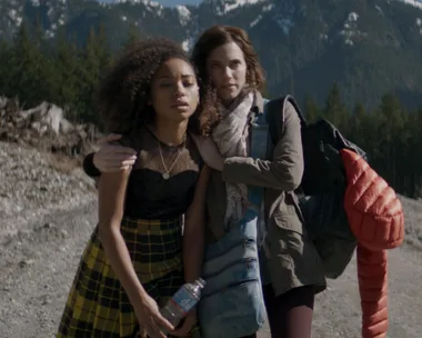 Two women walking on a mountain trail, one with curly hair holding a water bottle, and the other with a scarf and backpack.