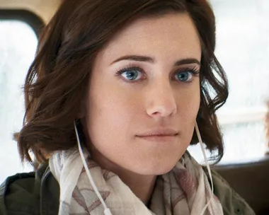 A woman with short brown hair and blue eyes listens to music on earphones, wearing a scarf and jacket, looking ahead.