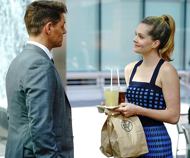 Man in a suit and woman in a blue dress holding drinks and bags, smiling at each other outdoors.