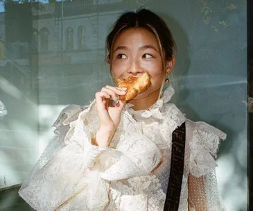 Woman in a lace blouse smiling while eating a pastry, standing in front of a storefront window.