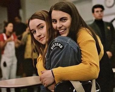 Two young women embrace in a classroom, one wearing a denim jacket with "SISTERS" text, both smiling warmly.