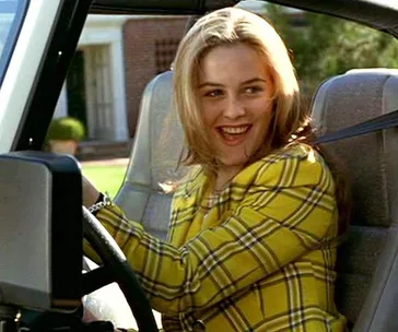 A woman in a yellow plaid jacket smiles while sitting in a car driver's seat.