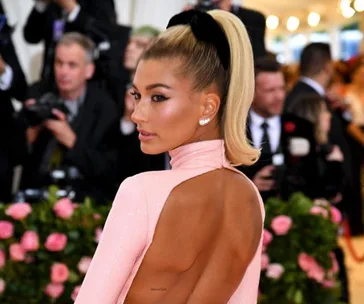 Woman in a pink open-back dress with a high ponytail at the 2019 Met Gala, surrounded by photographers and flowers.