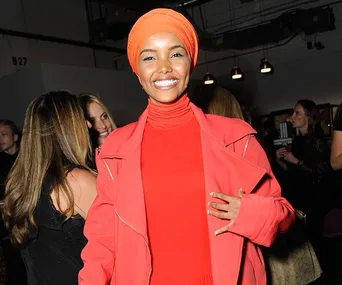 Halima Aden, smiling, wears an orange outfit and headscarf at an indoor event with dim lighting.