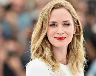 A close-up of Emily Blunt smiling, with light wavy hair and red lipstick, wearing a white top. The background is blurred.