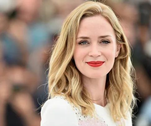 A close-up of Emily Blunt smiling, with light wavy hair and red lipstick, wearing a white top. The background is blurred.