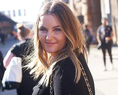 Woman with long blonde hair smiles outdoors during Fashion Week, wearing a black top and chain-strap bag.