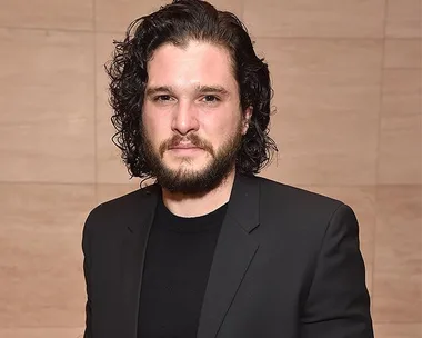 Man with curly hair and beard wearing a black suit, standing indoors.