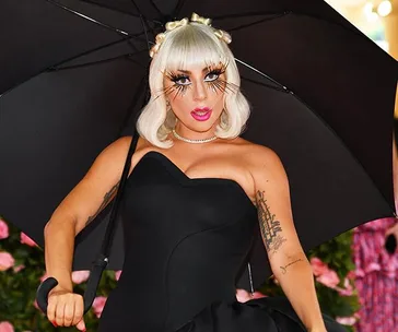 Lady Gaga at the 2019 Met Gala in a black dress holding a black umbrella, with bold makeup and statement lashes.