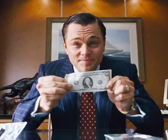 Person in a suit holding a dollar bill, seated at a desk with a painting in the background.