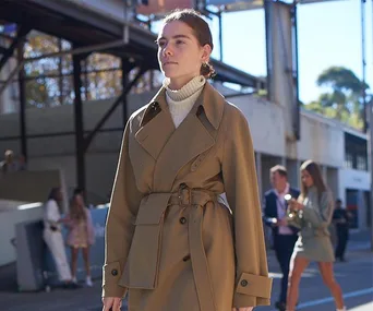 Person wearing a tan trench coat and turtleneck sweater walks outdoors during fashion week event.