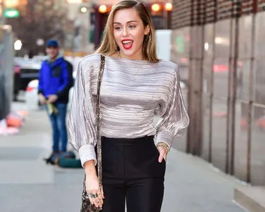 Woman in metallic top and black pants walking on a city sidewalk, smiling with red lipstick.