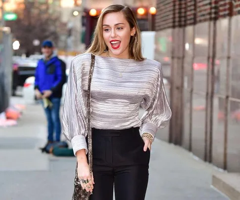 Woman in metallic top and black pants walking on a city sidewalk, smiling with red lipstick.