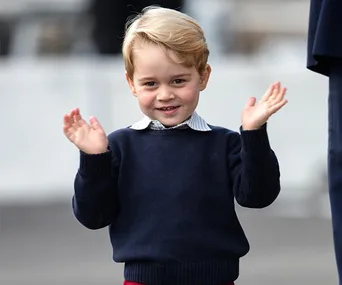 Young boy with blonde hair wearing a navy sweater, smiling with hands raised in a clapping motion.