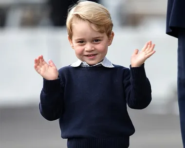 Young boy with blonde hair wearing a navy sweater, smiling with hands raised in a clapping motion.