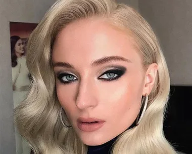 Blonde woman with smoky eye makeup, hoop earrings, and wavy hair poses indoors.