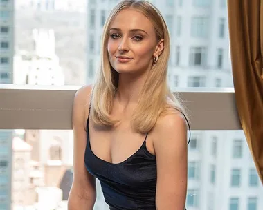 A person with long blond hair wearing a dark dress, sitting indoors with a cityscape view background.