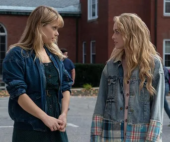 Two young women in denim jackets stand facing each other, mid-conversation, with a building in the background.