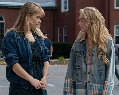 Two young women in denim jackets stand facing each other, mid-conversation, with a building in the background.