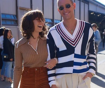 A smiling pair in stylish outfits, woman in brown top and man in a striped sweater, walking together outside a building.