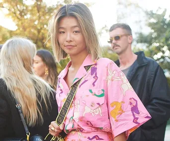 Young woman in a colorful pink outfit stands confidently outdoors at Sydney Fashion Week 2019, surrounded by people.