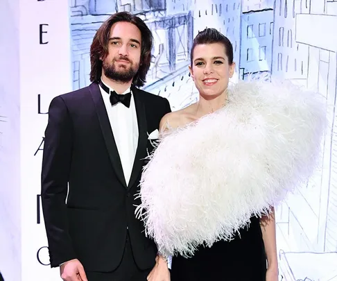 A couple in formal attire, the woman wearing a one-shoulder feather dress, smiling at an event with a city backdrop.