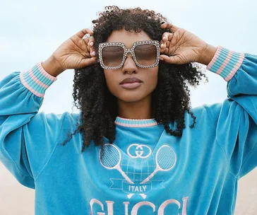 Woman in blue Gucci sweatshirt and rhinestone sunglasses on beach, adjusting frames with both hands.