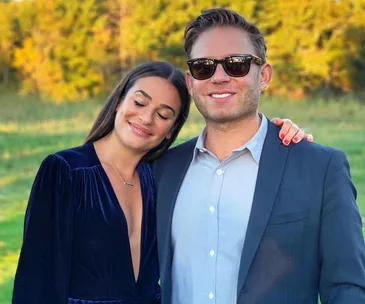 A couple poses outdoors; the woman in a blue dress leans on the man, who wears a suit and sunglasses, in a sunlit field.