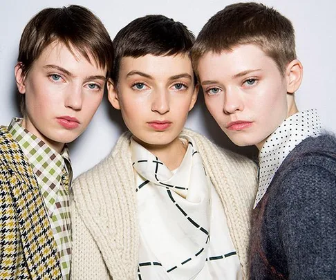 Three individuals with short hair and neutral expressions, wearing patterned shirts and sweaters, posing together indoors.