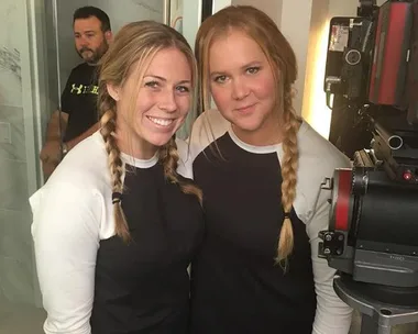 Two women with braided hair in matching outfits stand next to a camera on set, smiling.