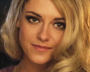 A woman with blonde hair and smoky eye makeup, smiling softly in a close-up shot.