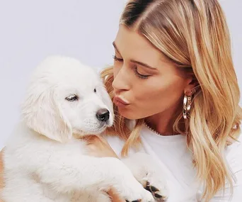 A woman with blonde hair kisses a fluffy white puppy she's holding close, both are in a bright setting.