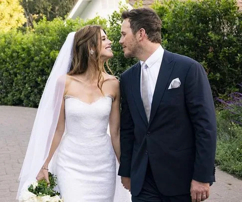 Bride in white dress and veil with groom in navy suit smiling at each other, walking outdoors holding hands.