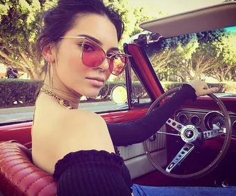 Woman in sunglasses sits in a red vintage car, holding the steering wheel, with trees in the background.