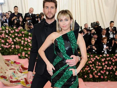 A man in a black suit and a woman in a green striped dress pose on a pink carpet surrounded by people in formal wear.