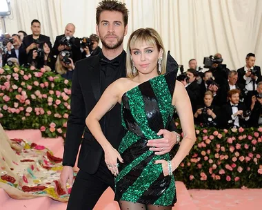 A man in a black suit and a woman in a green striped dress pose on a pink carpet surrounded by people in formal wear.