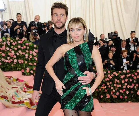 A man in a black suit and a woman in a green striped dress pose on a pink carpet surrounded by people in formal wear.