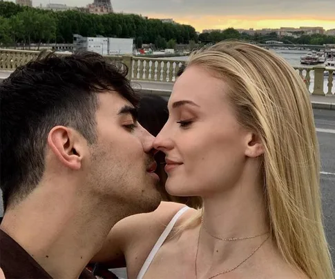 A couple sharing a close, intimate moment on a bridge, with scenic views of the river and cityscape in the background.