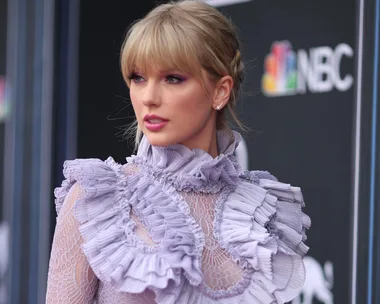 A woman in a lavender ruffled dress poses in front of a backdrop with the NBC logo.