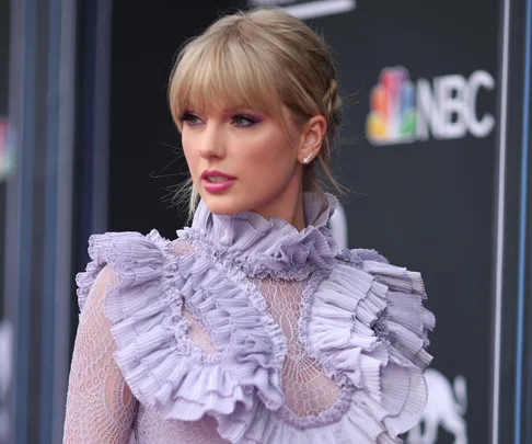 A woman in a lavender ruffled dress poses in front of a backdrop with the NBC logo.